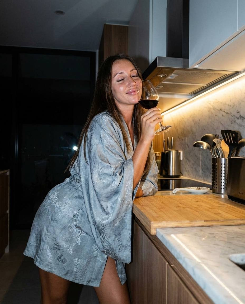 A girl with long straight hair leans against a wooden kitchen unit, holding a glass of red wine to her face, eyes closed.