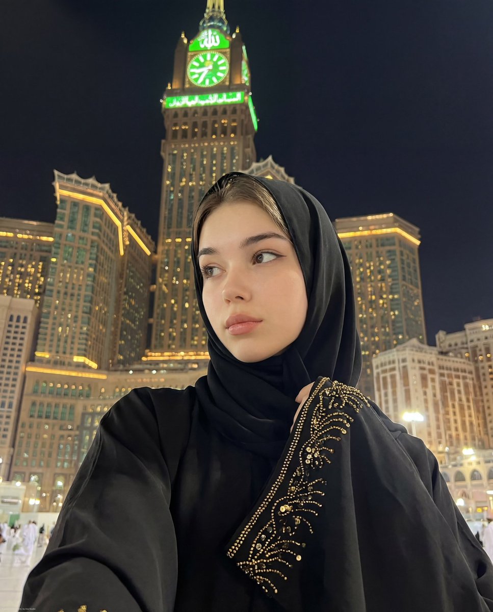 A serene young woman in a black abaya and hijab, standing near the illuminated Makkah Royal Clock Tower, captured in a close-up night selfie.