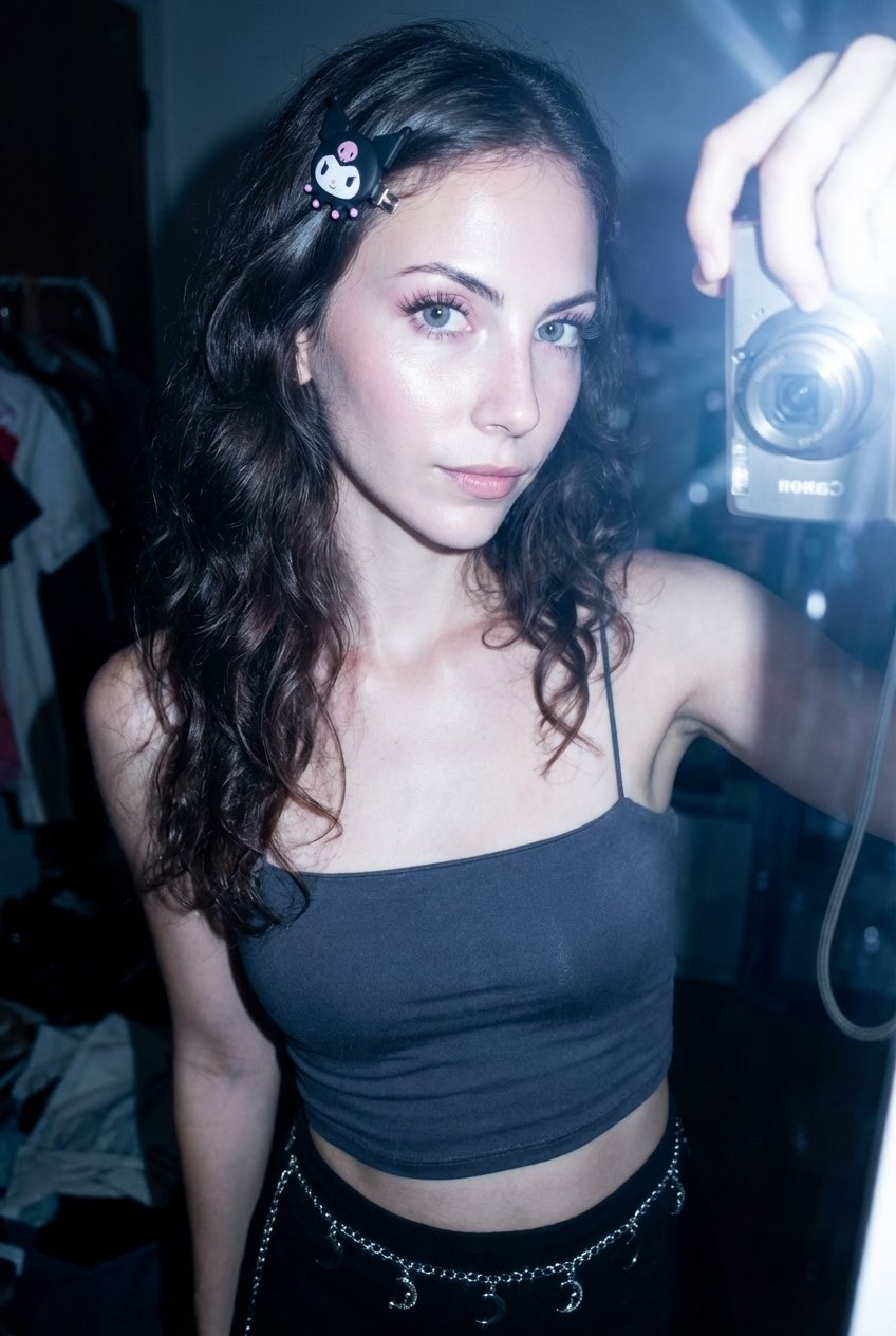 A teenage girl with dark brown wavy hair wearing a dark grey crop top, posing in front of a mirror, showcasing bright eyes and radiant skin.