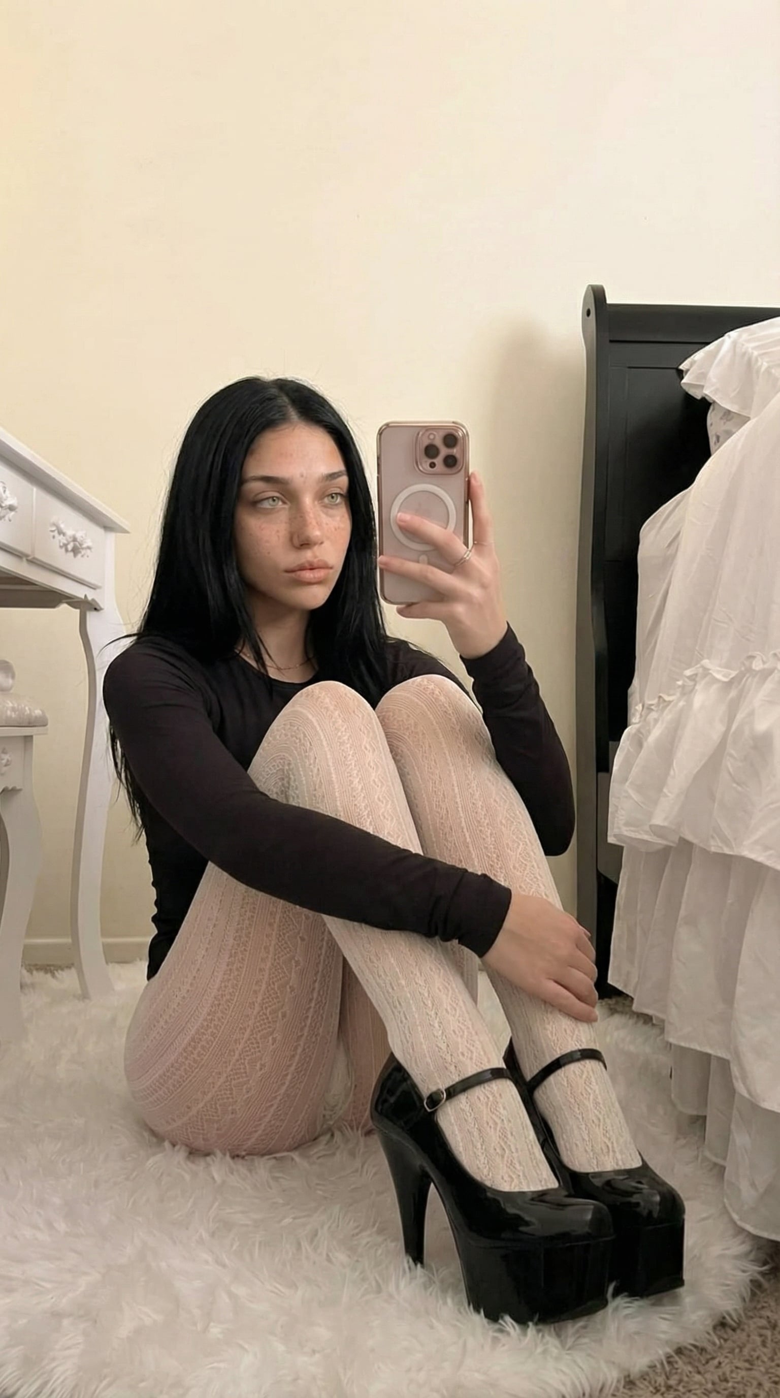 A woman in black lace tights and platform heels takes a selfie on a plush white rug in a softly lit bedroom.
