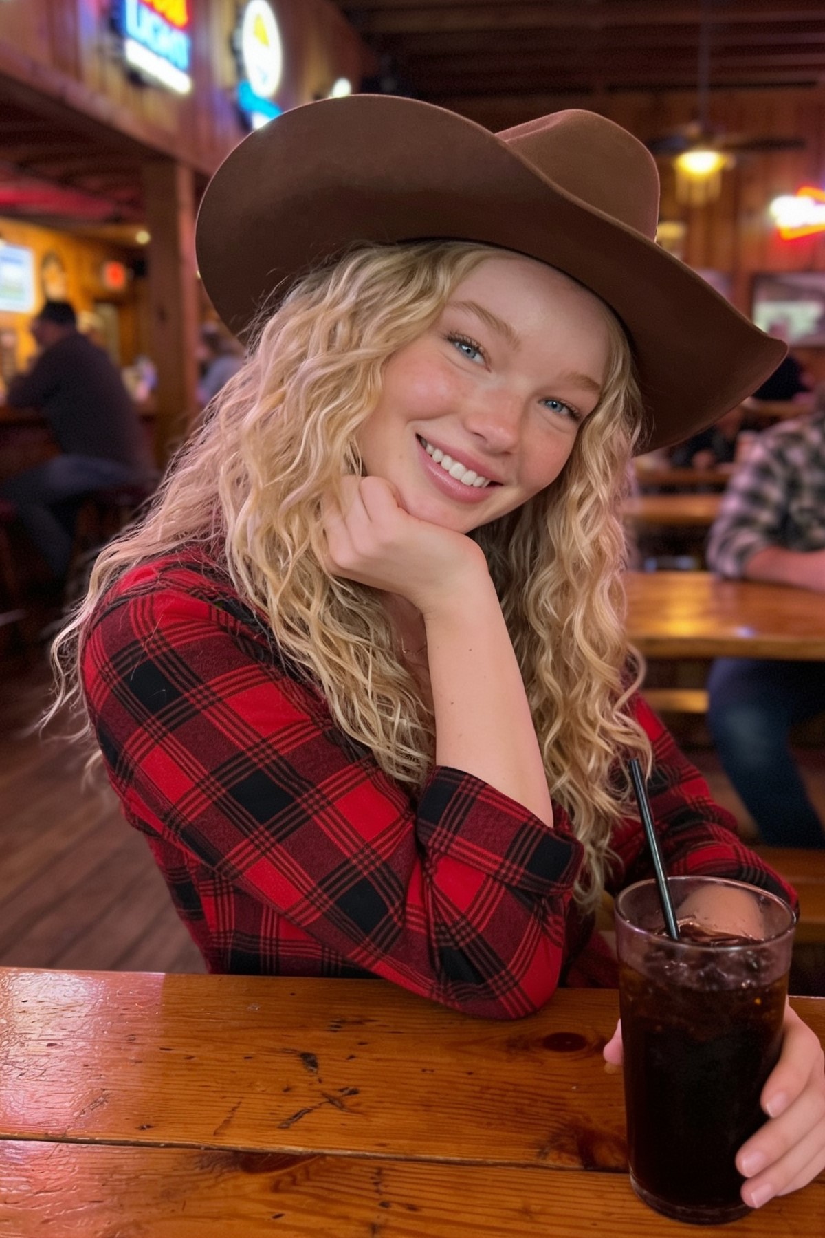 Close-up portrait of a person in a brown cowboy hat and plaid shirt, smiling at a rustic bar with a glass of soda