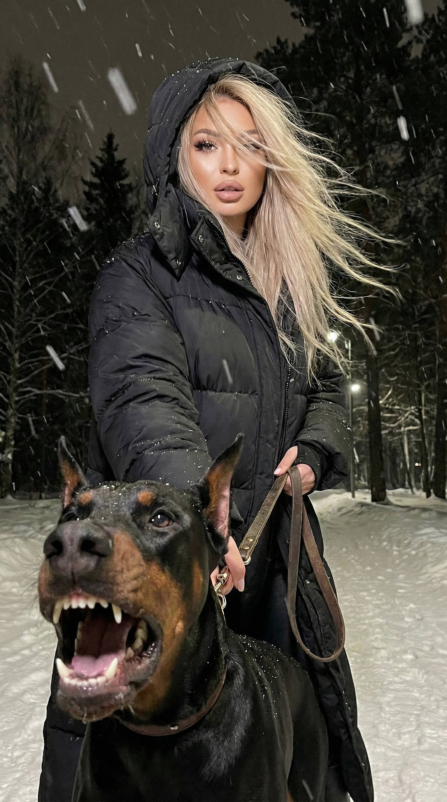 A girl in a matte puffer jacket posing in a snowy forest with a Doberman barking in the foreground.