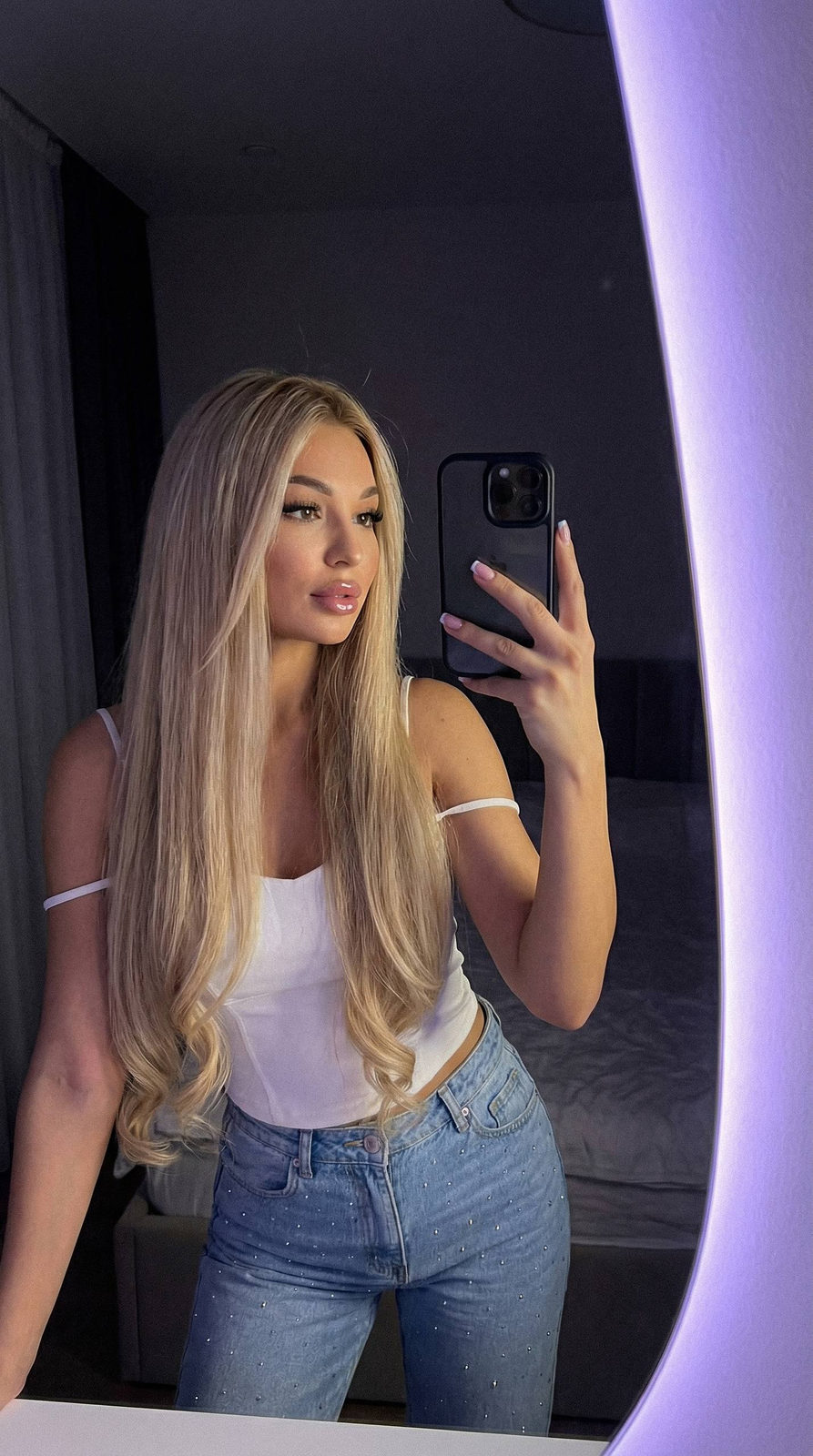 A woman taking a selfie in a mirror with purple lighting, holding a black phone, wearing a white top and embellished light blue jeans.