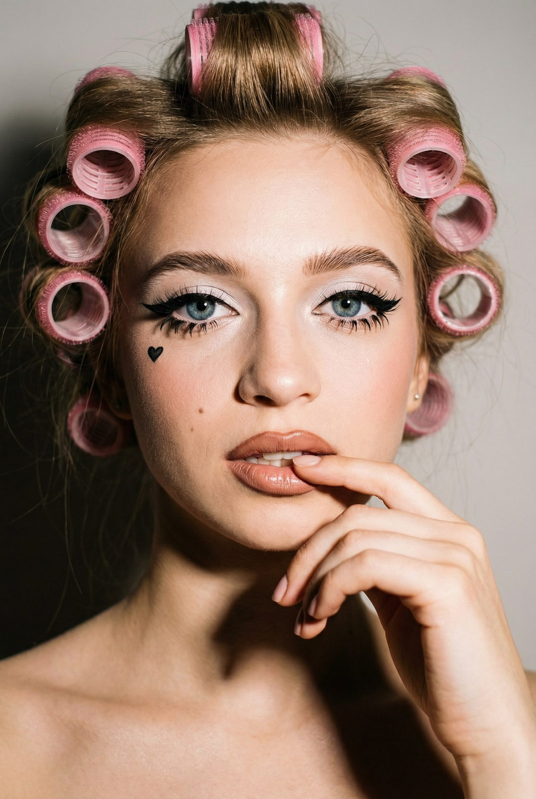 Close-up portrait of a woman with pink hair rollers, wearing doll-like makeup and a playful expression.