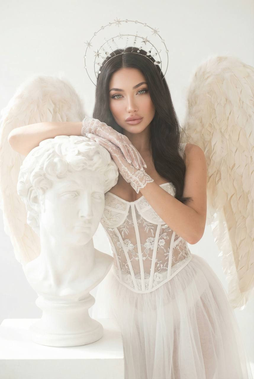 A model in an intricate corset dress with floral embroidery, flanked by large angel wings, gently resting on a classical bust in a luminous studio.