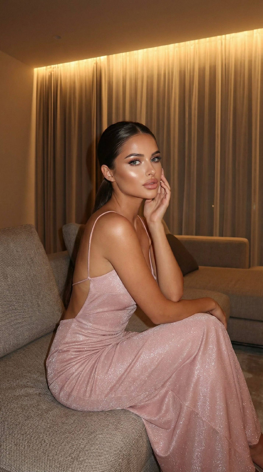 A woman in a soft pink shimmer dress sits gracefully on a modern lounge sofa in a cozy night interior with warm lighting.