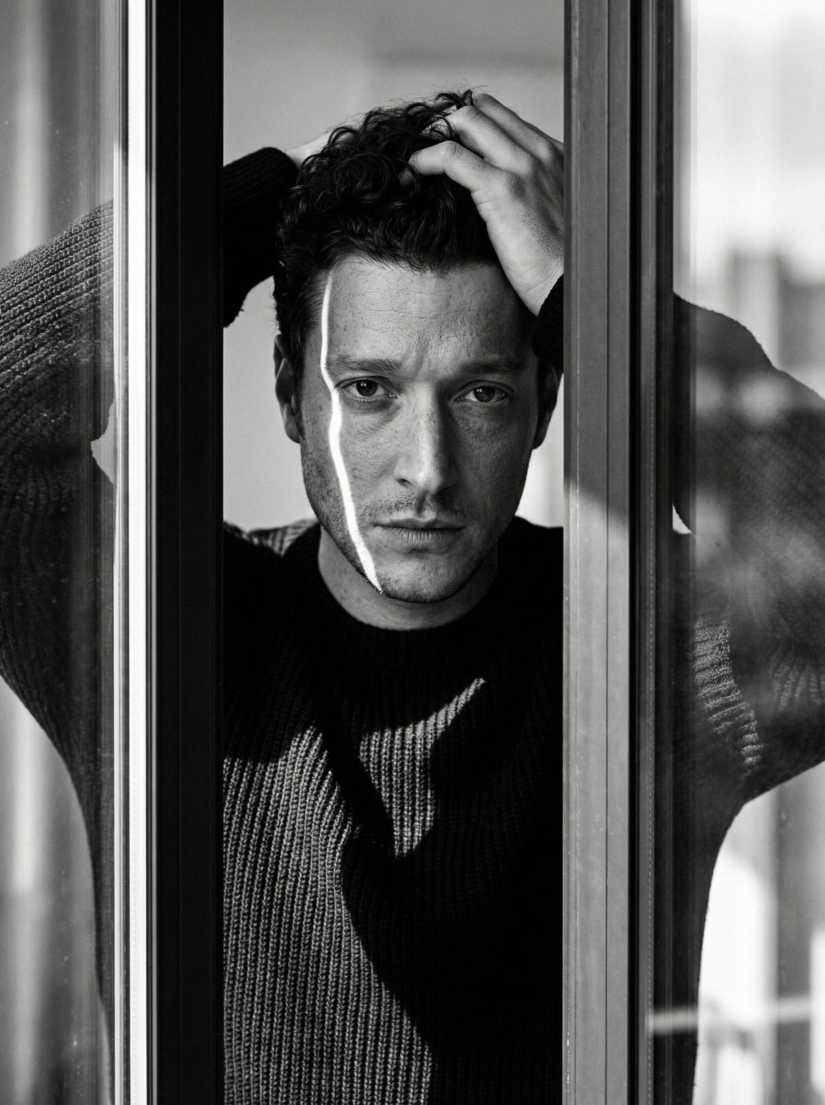 A man in a charcoal sweater stands in a dark window frame, gazing at the camera with a relaxed expression and partially obscured body.