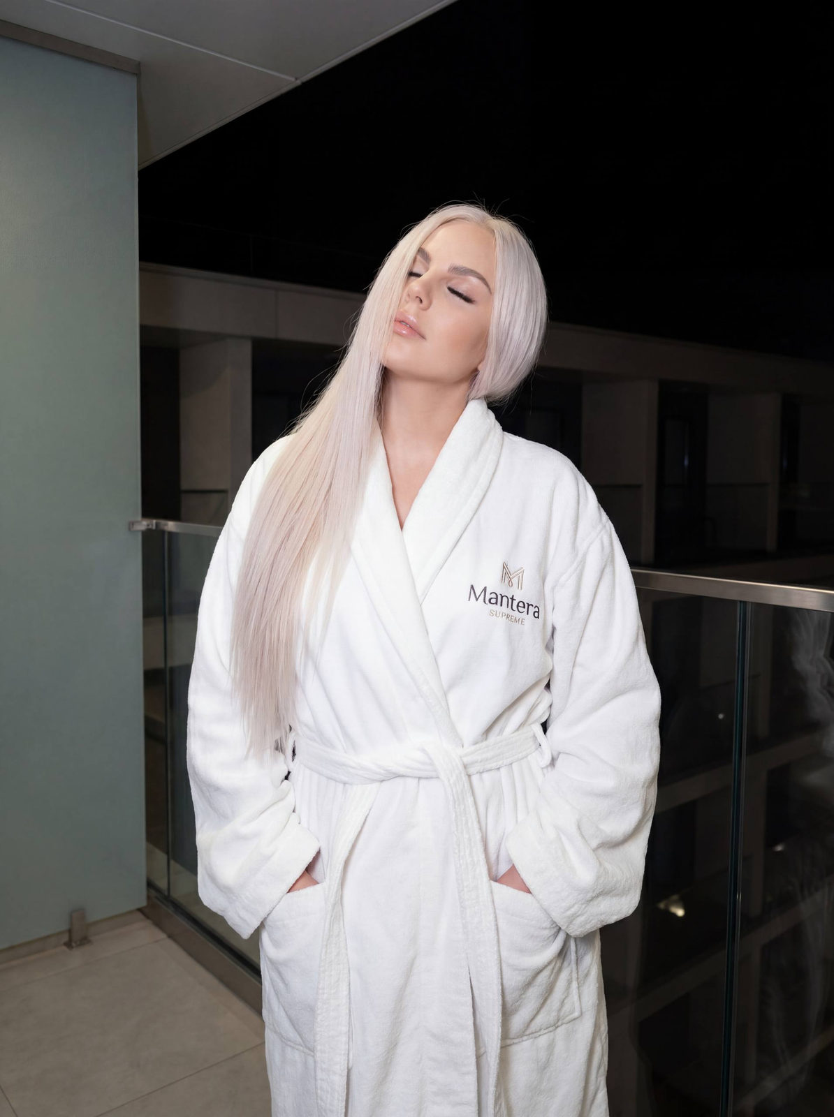 A girl in a white oversized bathrobe stands on a minimalist balcony with glass railing, eyes closed and hair styled to one side.