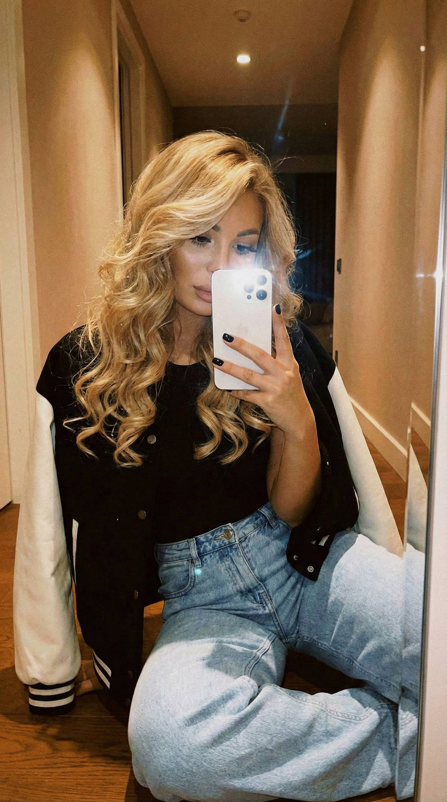 A woman takes a selfie in front of a mirror, wearing a black top and light blue high-waisted jeans, with a monochrome bomber jacket draped over her shoulders.