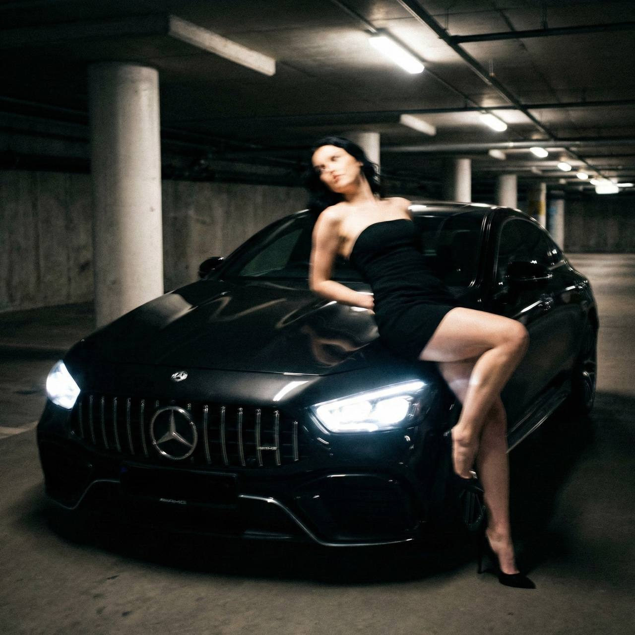 A woman reclining on a Mercedes-Benz AMG GT 43 in a dark garage, wearing a strapless dress, captured with motion blur and reflections.
