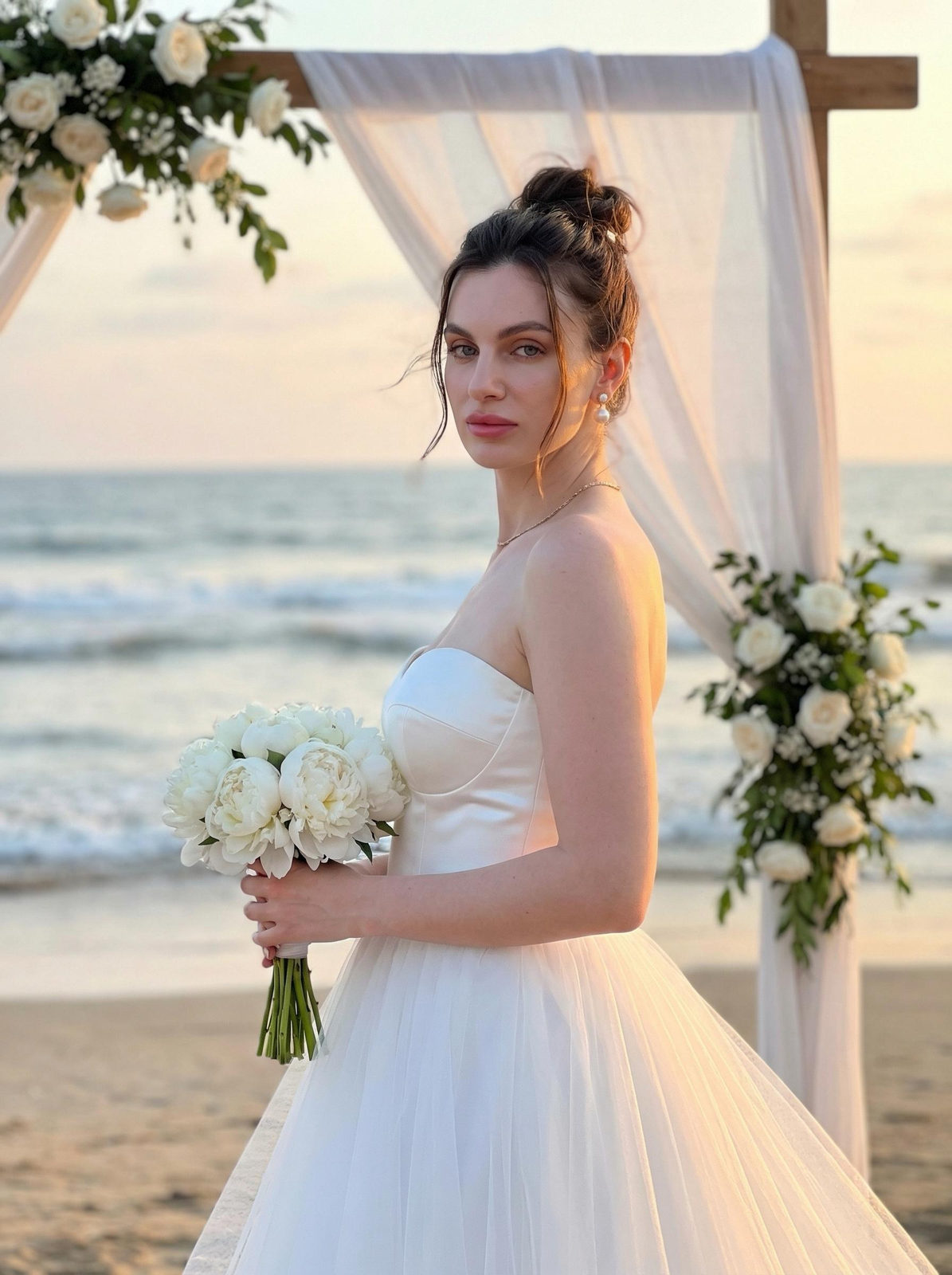 A bride in a white wedding dress stands by a floral arch on the beach, holding a bouquet of white peonies, with a sunset backdrop.