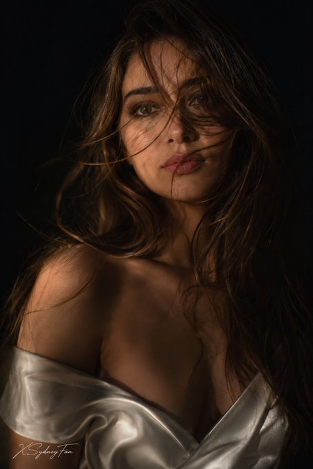 Intimate close-up of a woman with windswept dark hair dressed in a white satin garment against a deep black background.