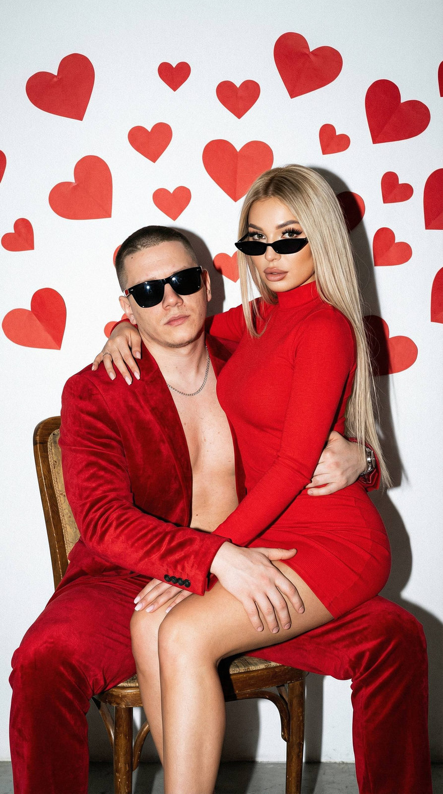 A couple poses against a white wall, the man in a red velvet suit and the woman in a matching red dress, both exuding confidence.