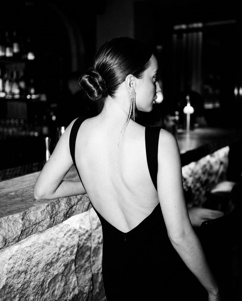 A woman in a backless evening dress, showcasing her back and a long diamond earring, set in a dimly lit luxury lounge.