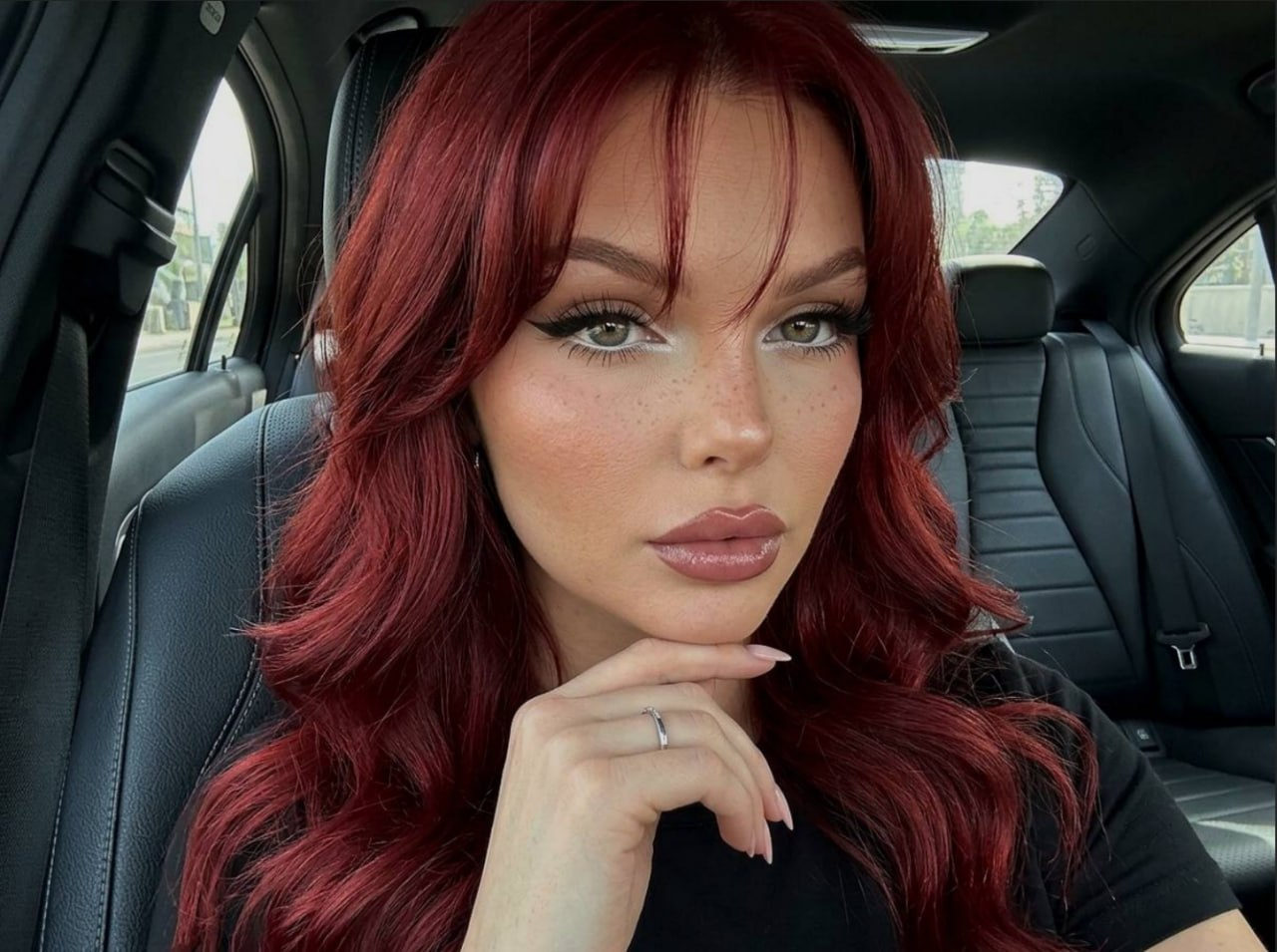 Close-up selfie of a woman in a car, calm expression with glossy hair and soft waves, wearing black, softly illuminated by diffused daylight.