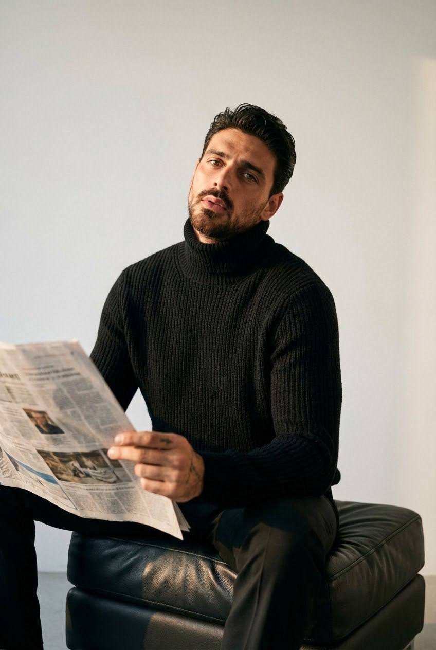 A man in a bulky black turtleneck reading a newspaper while sitting on a black leather pouf, captured in soft golden hour lighting.