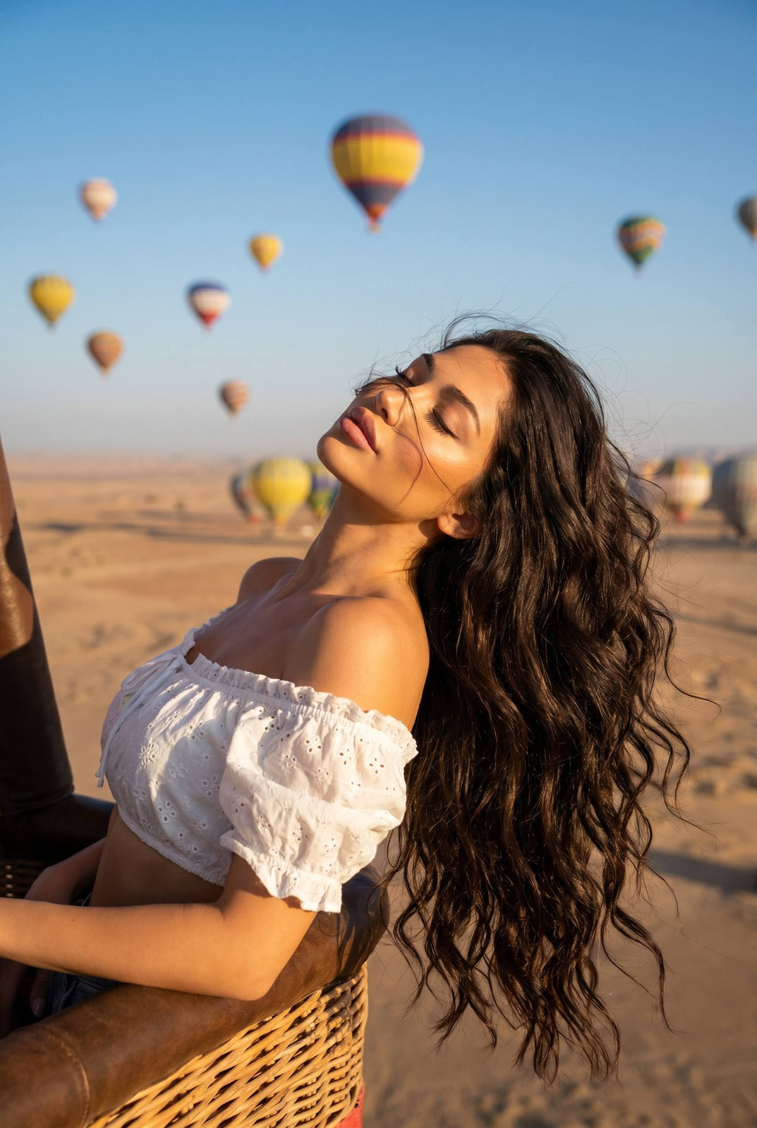 A woman in a white crop top relaxing in a hot air balloon, golden hour sunlight illuminating her face and hair with colorful balloons in the background.