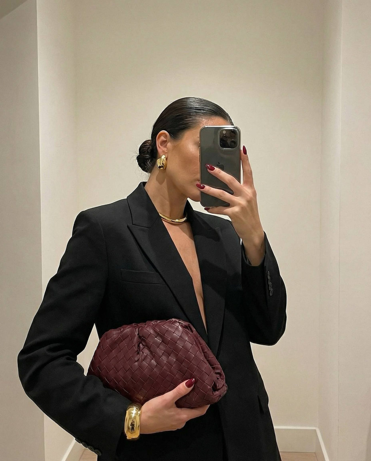 A stylish mirror selfie showcasing a woman in a black blazer with a burgundy clutch, elegant gold accessories, and a calm expression.