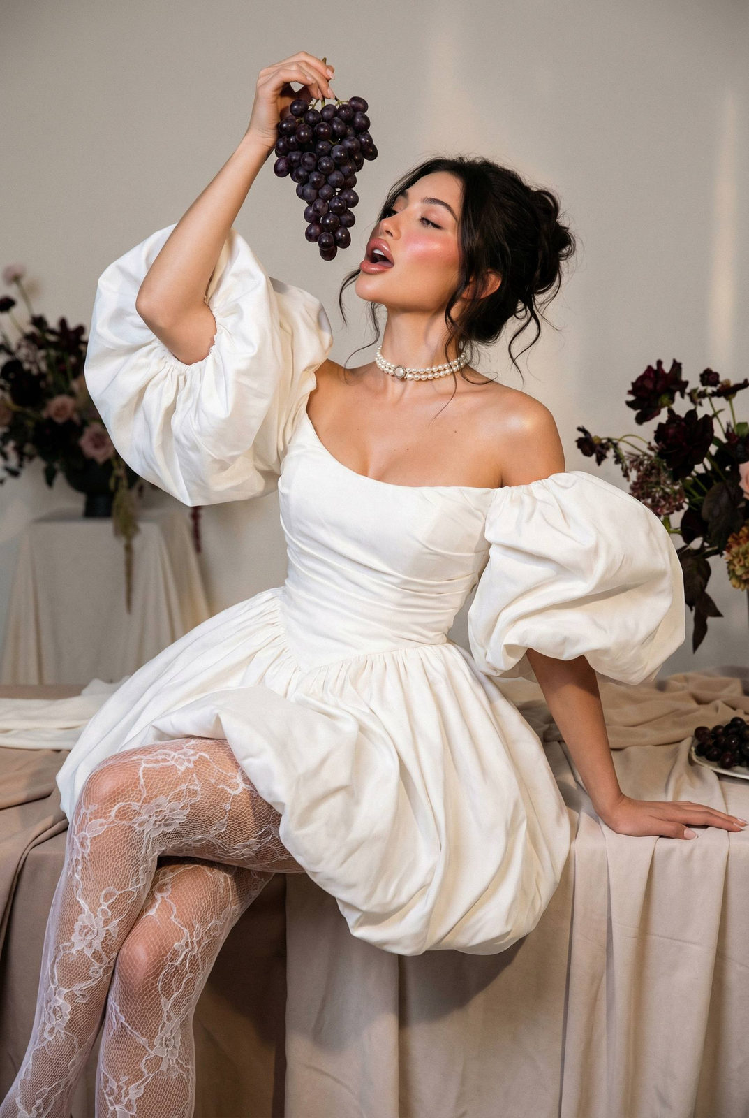 High-fashion portrait of an elegant woman poses gracefully with dark grapes, wearing a structured white mini dress and lace tights.