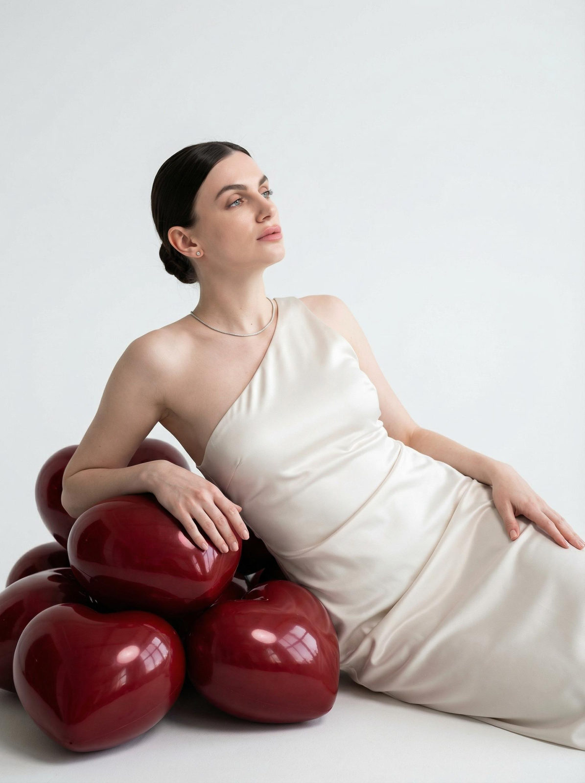 A woman in a flowing one-shoulder dress poses with glossy heart-shaped spheres, soft lighting enhances her elegant look.