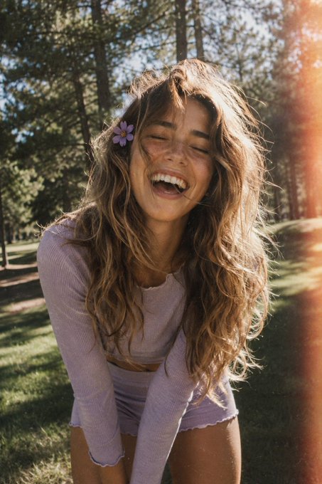 Woman in lavender two-piece laughing in a sunlit park, with a flower behind her ear.