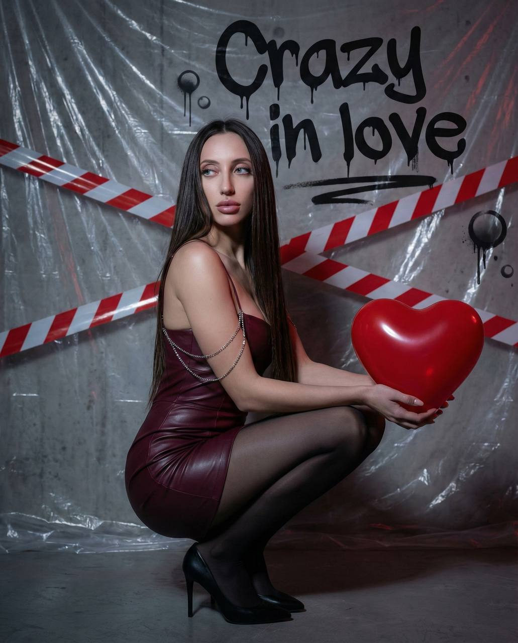 A woman in a burgundy leather dress squats holding a shiny red heart, with a dramatic concrete wall backdrop and soft diffused lighting.