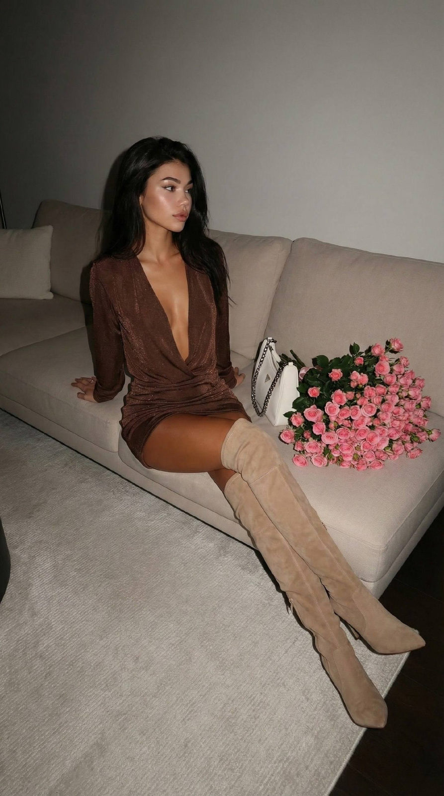A woman in a shiny brown top and mini skirt lounges on a light sofa, with soft lighting creating depth and shadows.