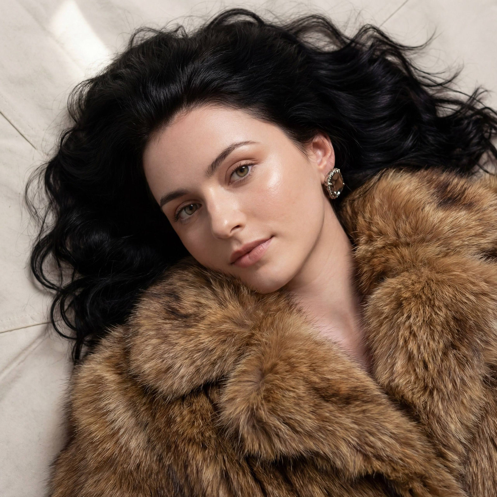 A serene woman lying down with soft radiant skin and voluminous brown fur jacket, gazing thoughtfully towards the camera.