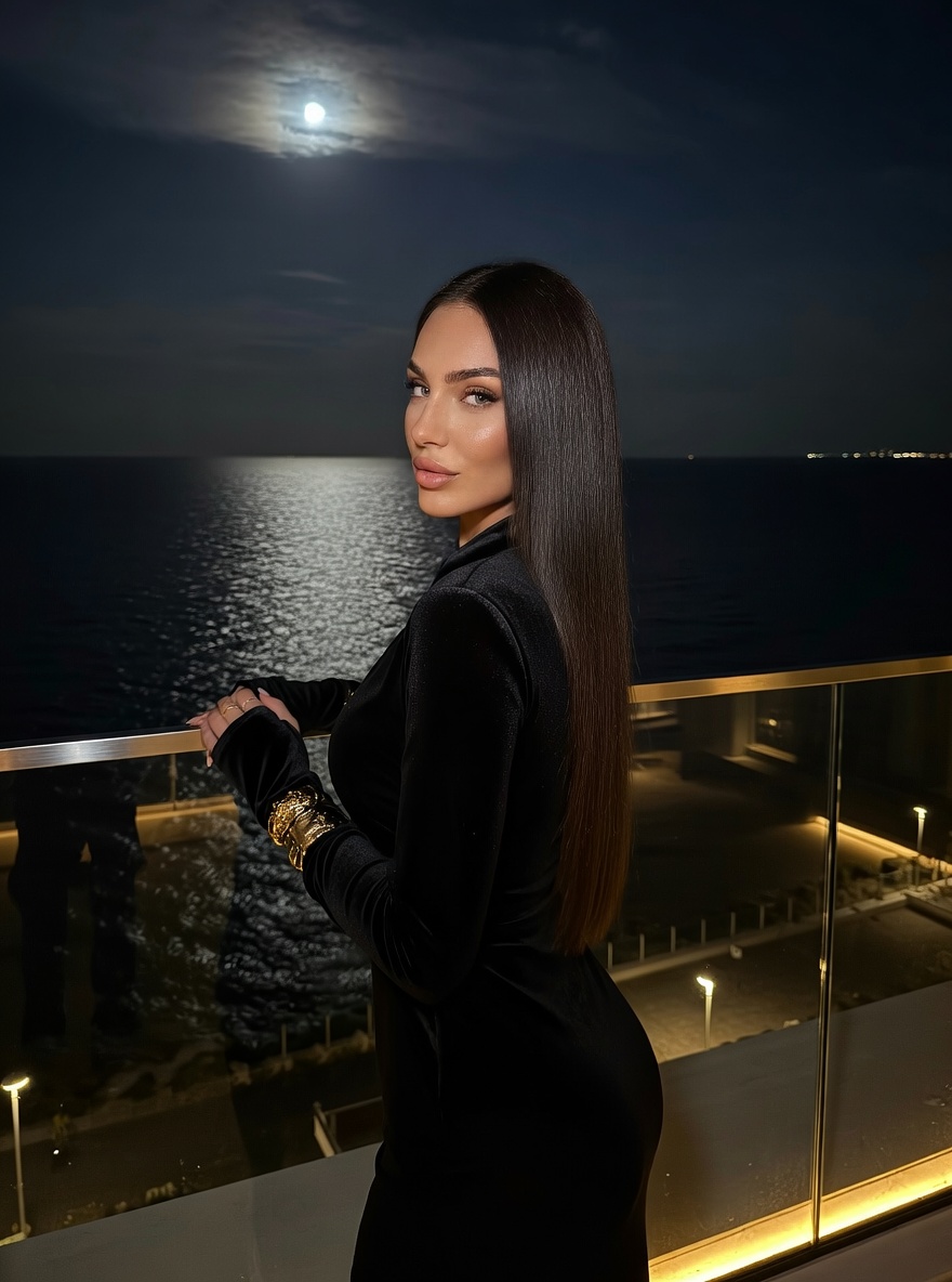 A woman in a matte black dress stands by a glass fence at night, illuminated by moonlight and hotel lights.