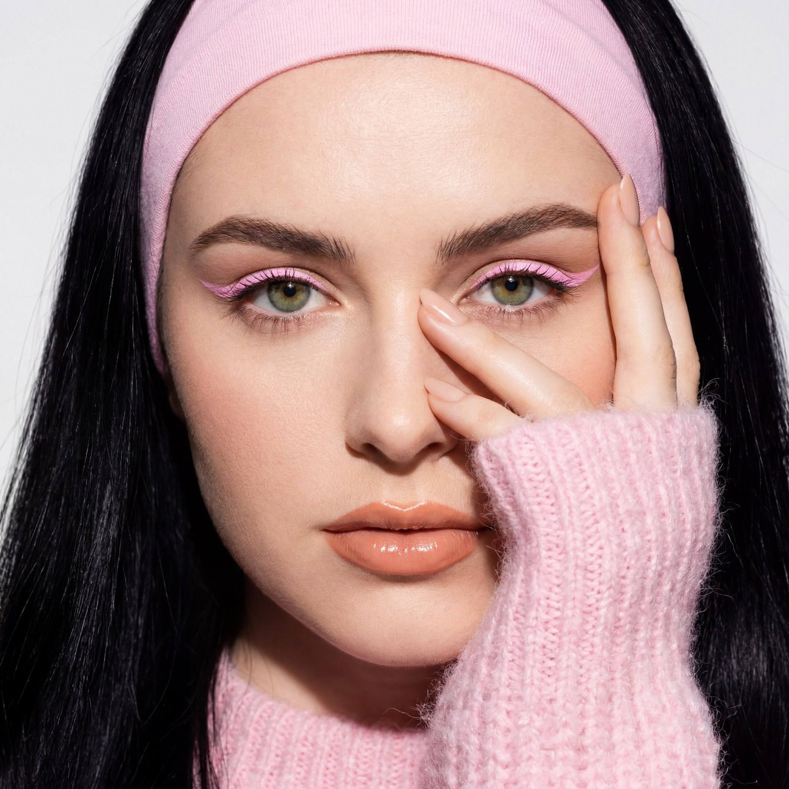 A woman in a fluffy pink sweater, posing with a hand covering part of her face, showcasing impeccable makeup under studio lighting.
