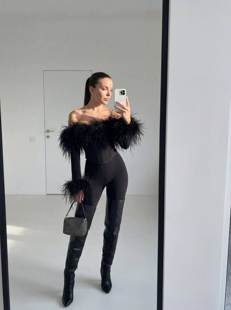 A woman in a fitted outfit with feather details, taking a selfie in a large mirror within a minimalist room, sunlight casting soft highlights on her face.