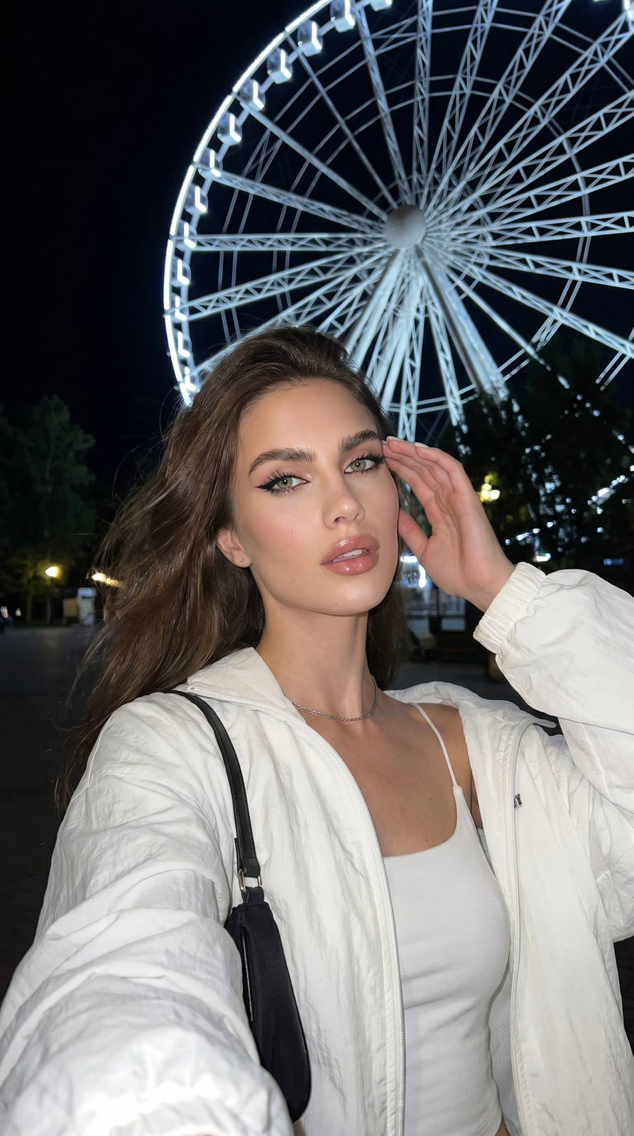 Close-up selfie of a woman in a white windbreaker, Ferris wheel lights in the background, vibrant night atmosphere.