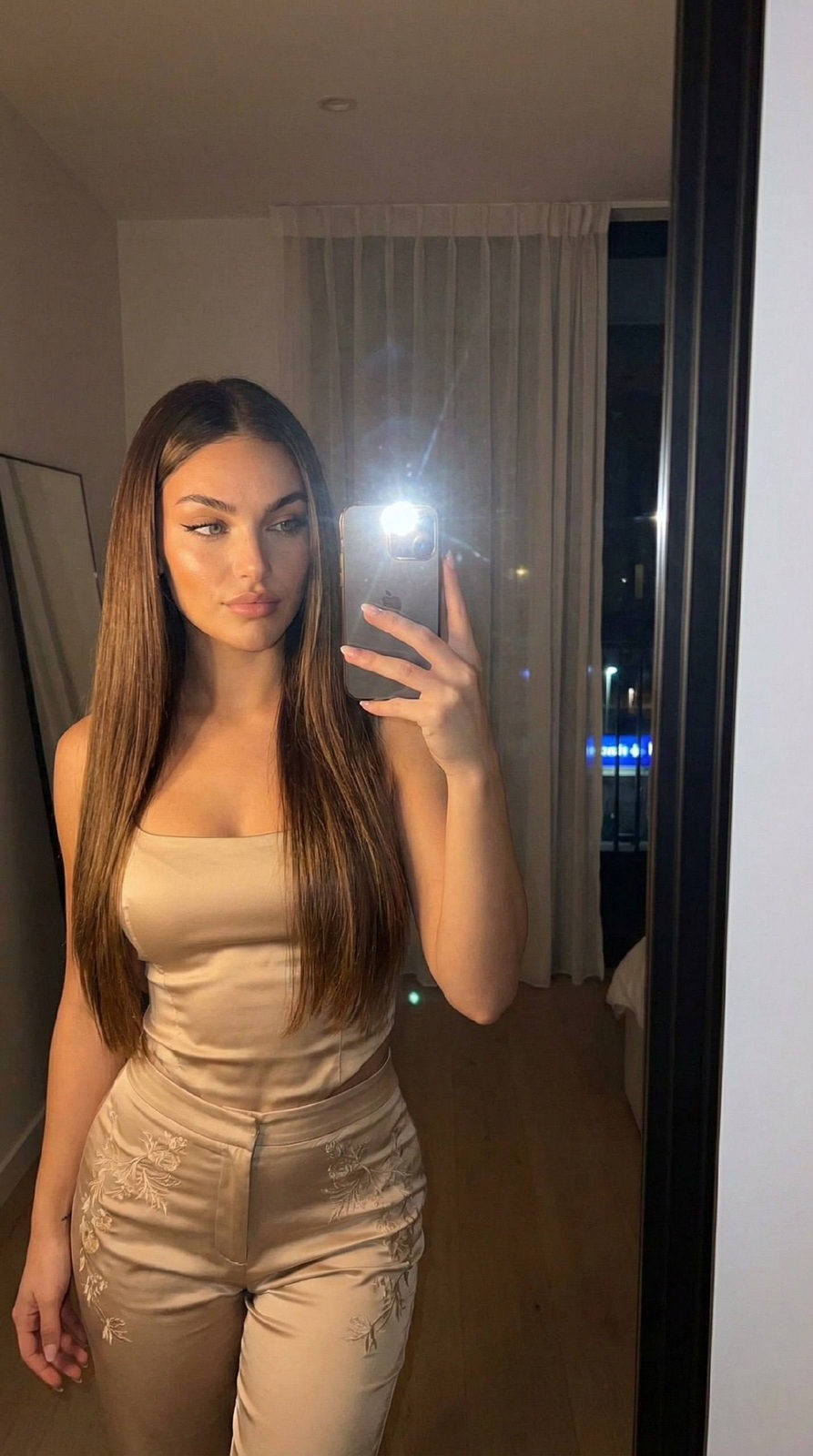 A young woman taking a mirror selfie, dressed in a stylish beige satin outfit with soft evening light creating gentle shadows.