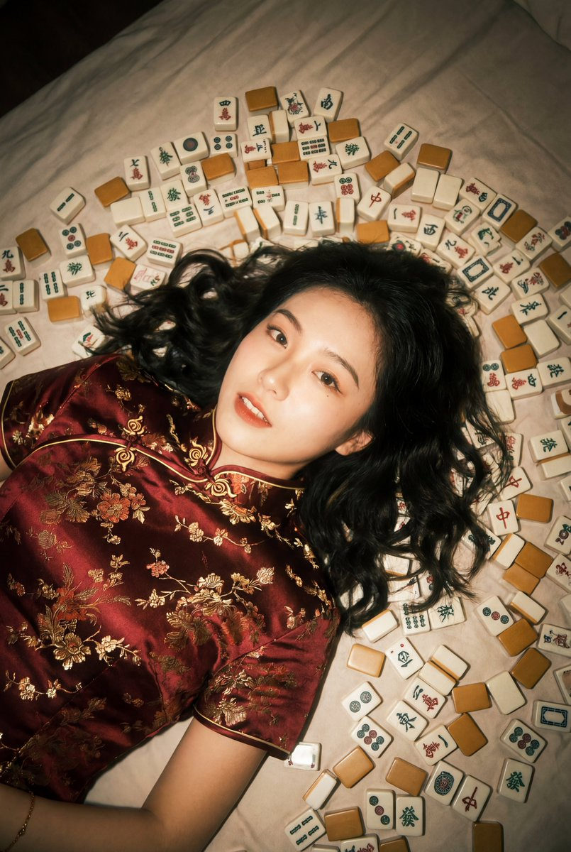 A cute young Chinese woman in a silk Qipao lying on a bed with Mahjong tiles, captured with a dreamy, cinematic aesthetic.