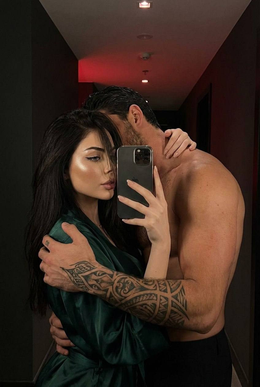 A close-up of a woman in a dark green silk robe, taking a mirror selfie, with a man hugging her from behind in a dark hallway.