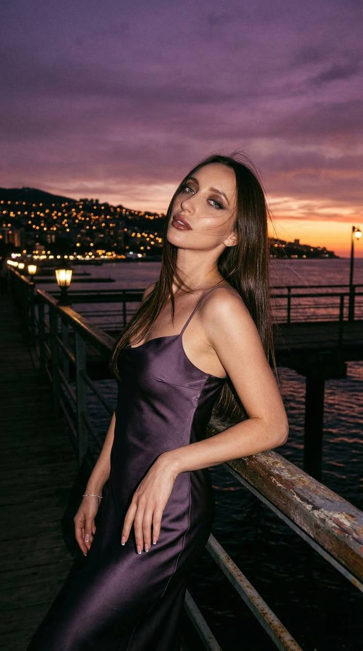 Cinematic portrait of a girl in a dark lavender silk dress, standing by a pier with city lights and a violet-pink sky.