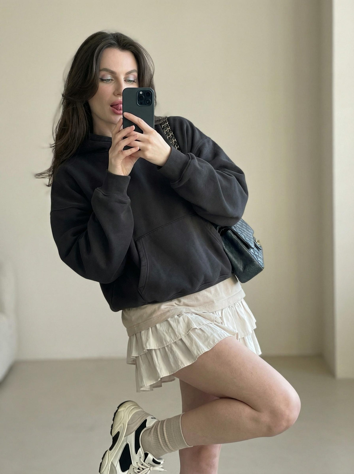 A woman taking a mirror selfie, sporting soft pastel eyeshadow and a relaxed pose in a minimalist interior.