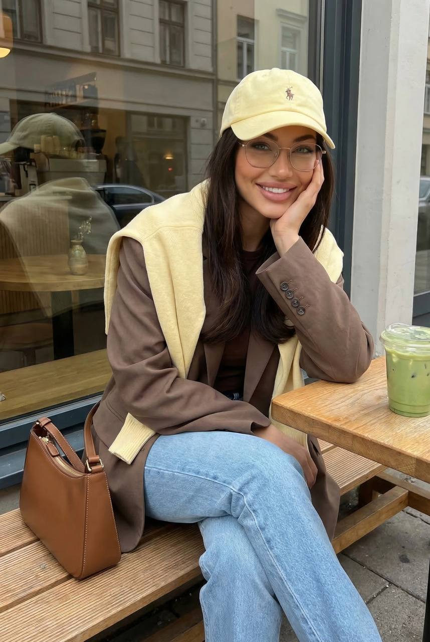 A woman in a brown blazer and yellow sweater sitting at an outdoor cafe table, smiling warmly, with a green iced drink beside her.