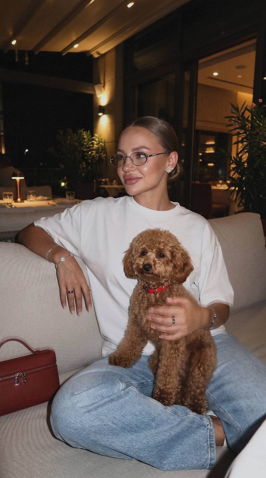 A girl on a bright sofa holds a caramel poodle, wearing Miu Miu glasses and casual attire, against a warm restaurant backdrop.