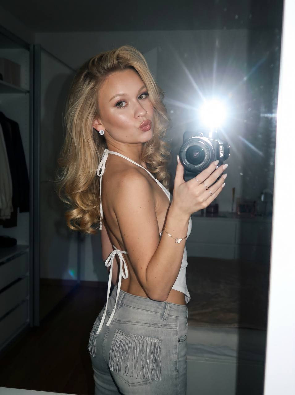 A beautiful girl posing confidently in front of a mirror, holding a Nikon camera, with camera flash creating natural light flares.