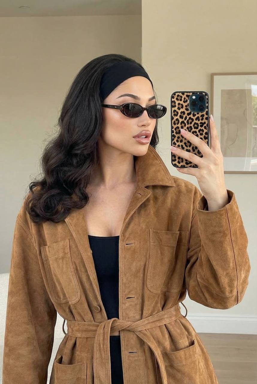 A woman in a camel jacket taking a mirror selfie, posed elegantly with her phone.