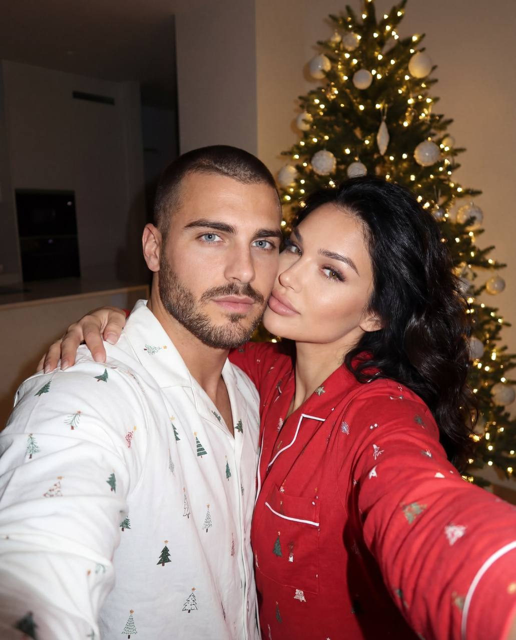 A couple in cozy pajamas taking a selfie in front of a Christmas tree with warm lights.