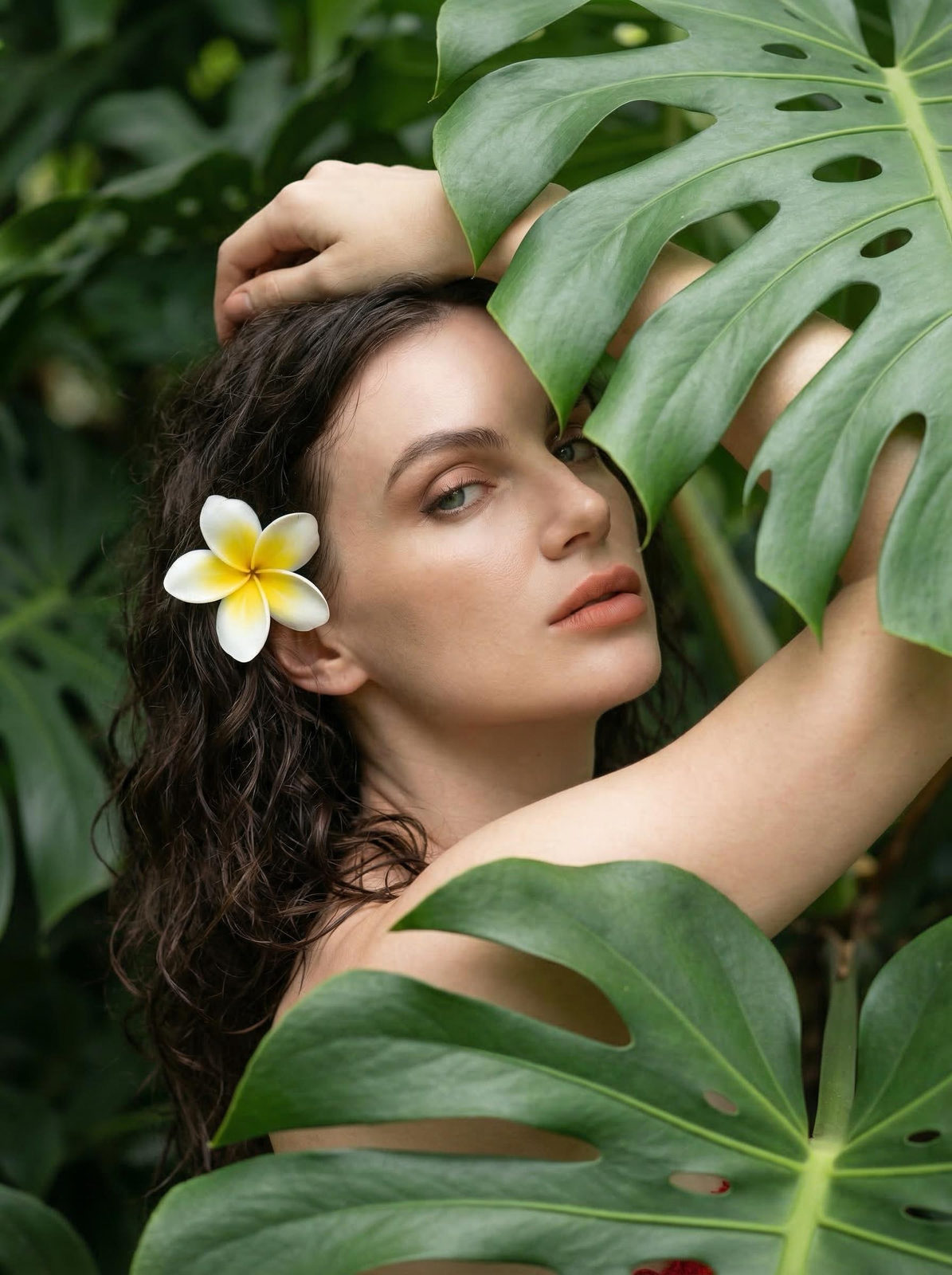 A woman surrounded by dense tropical vegetation, her head tilted back, gazing at the camera with soft, natural makeup and a frangipani in her curls.