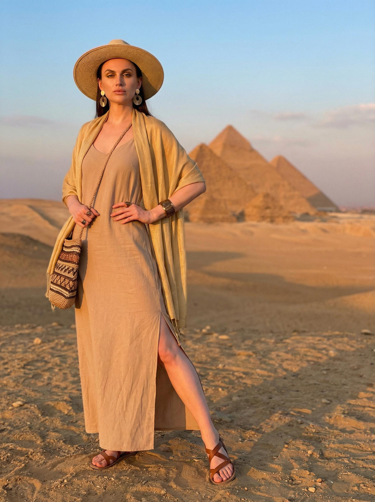 Woman in a long, sand-colored maxi dress standing in a desert with Egyptian pyramids behind her, wearing a straw hat and carrying a woven bag.