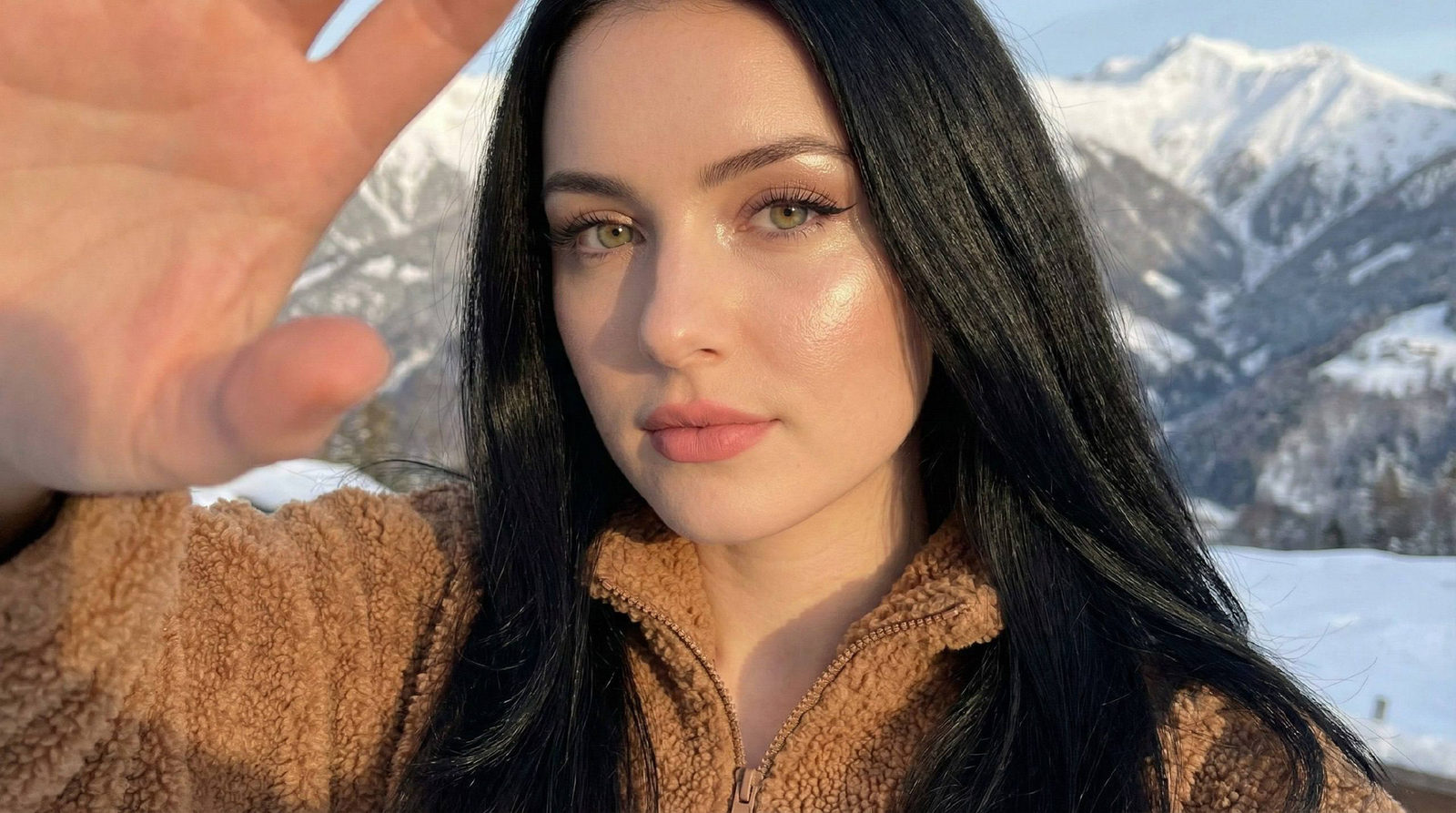 A sunlit winter selfie featuring a woman with long, glossy hair and a teddy zip-up sweater, posing with a warm smile against snowy mountains.