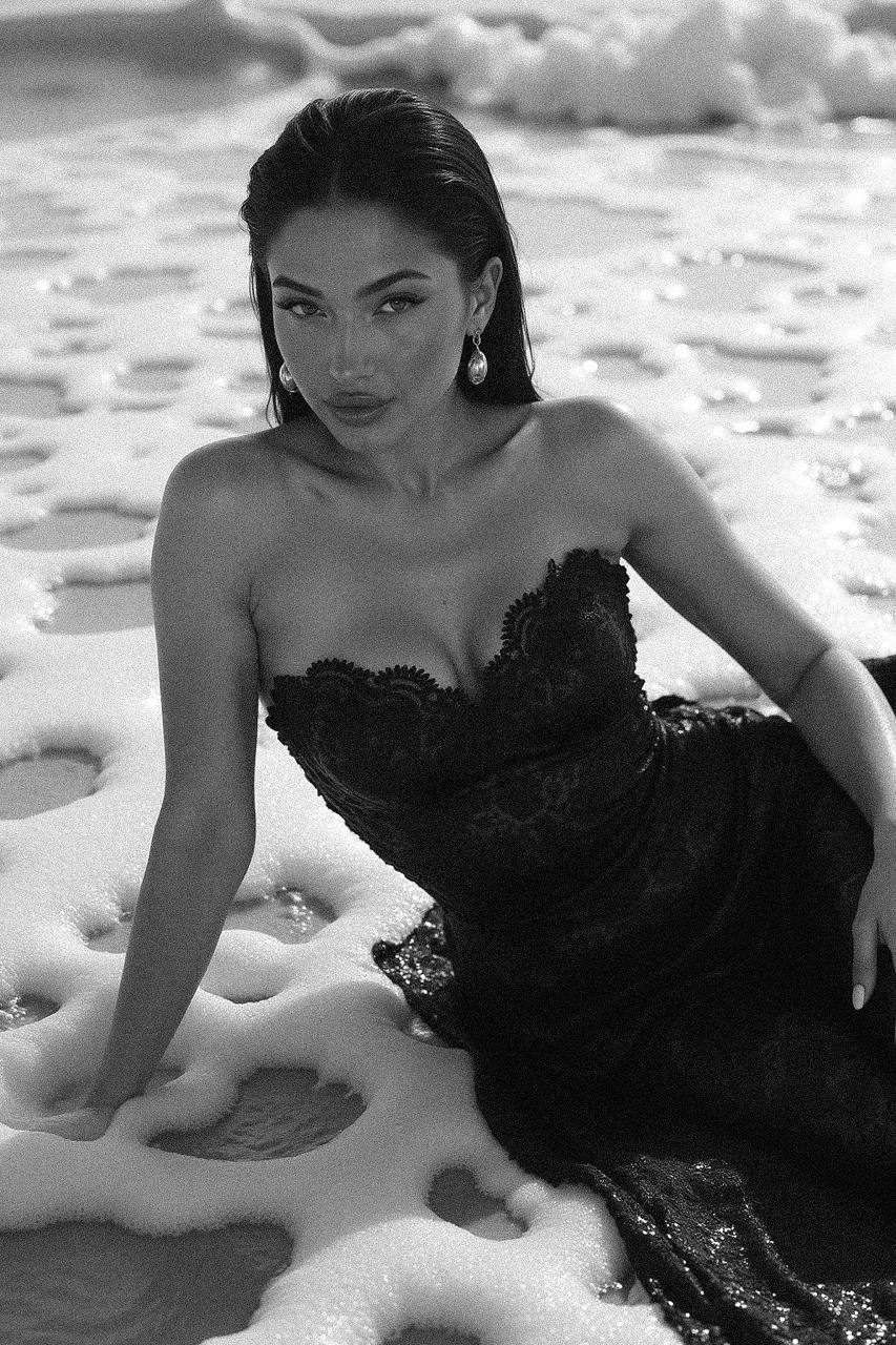 A woman in an elegant lace dress lies gracefully in shallow ocean waves, her pose exuding confidence while large statement earrings catch the light.