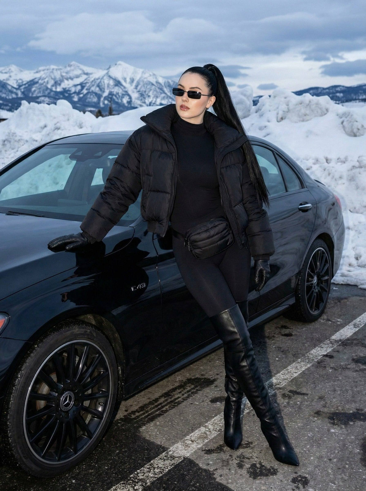 A stylish figure leans against a sleek black Mercedes Benz on a snowy parking lot, wearing a black bomber jacket and high-heeled boots.