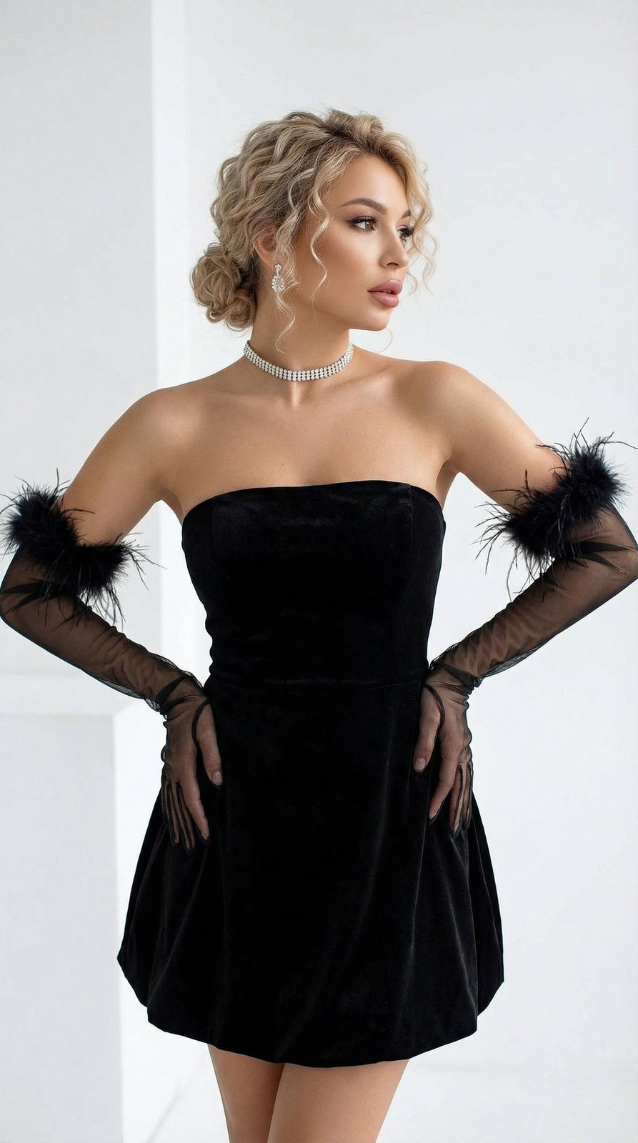 A young woman in a black velvet mini dress poses in a minimalist studio, with long black gloves and a confident expression.