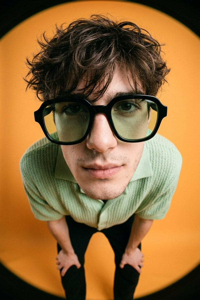 A man in a pastel green knitted shirt with oversized black-framed sunglasses, posing in a fisheye close-up against a solid orange background.