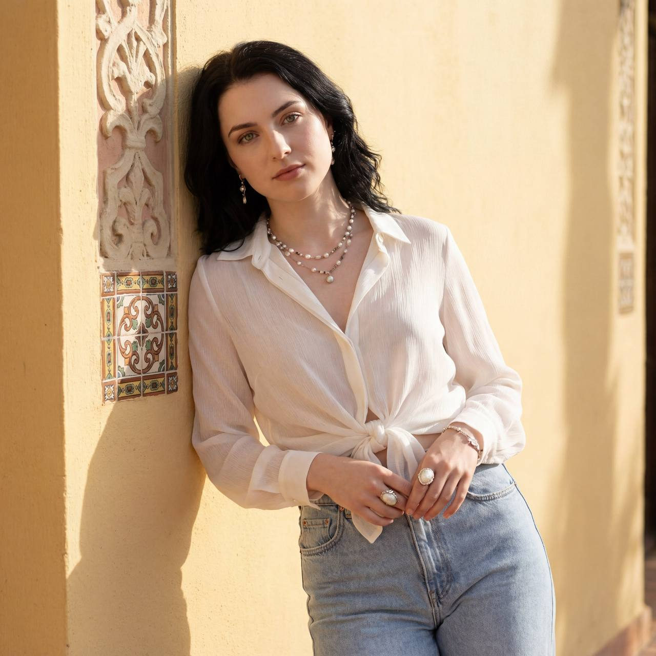 Woman with long dark wavy hair in a light blouse, leaning against a yellow wall, sunlit and relaxed.